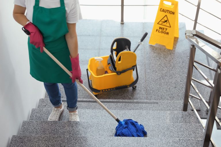 Person wearing a green apron and rubber gloves mopping a tiled staircase with a wet floor sign in the background