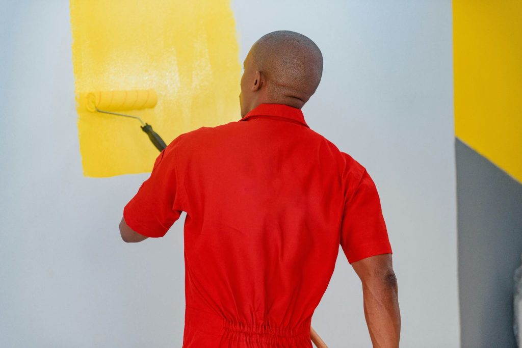 man in red overalls painting a grey wall yellow